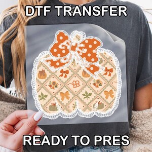 May include: A DTF transfer sheet featuring a pumpkin-shaped graphic with a white lace border. The pumpkin has a diamond pattern filled with small icons of pumpkins, sunflowers, leaves, and bows, topped with a large orange polka-dot bow.