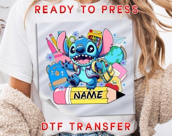 May include: A DTF transfer featuring a cartoon character, surrounded by school supplies. The character is blue with large ears and is holding a backpack. A yellow pencil with the word "NAME" is in the foreground. The words "READY TO PRESS" and "DTF TRANSFER" are in red.