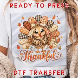 May include: A white t-shirt featuring a DTF transfer graphic of a cartoon turkey with large eyes, wearing a leopard print sweater and a plaid bow. The turkey is surrounded by autumn leaves and the word Thankful in orange script lettering.