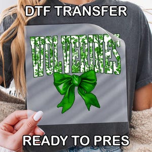 May include: A square DTF transfer sheet featuring the word WOLVERINES in a green and white leafy pattern, positioned above a large, bright green decorative bow. The design is printed on a clear film, ready for heat application onto fabric.