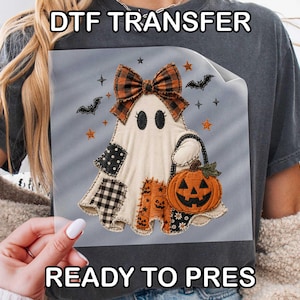 May include: A DTF transfer sheet featuring a ghost graphic with a plaid bow, holding a jack-o'-lantern pumpkin. The design includes black and white patterned patches on the ghost, small bats, and stars. The text reads DTF TRANSFER READY TO PRES.