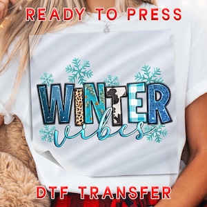 May include: A white t-shirt featuring a graphic design with the text "WINTER vibes" in blue, leopard print, and cow print patterns. The design is accented with light blue snowflake illustrations and is labeled as a ready to press DTF transfer.