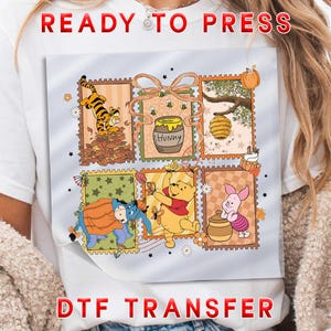 May include: White t-shirt featuring a DTF transfer with a colorful design of Winnie the Pooh characters in a postage stamp style. The words "READY TO PRESS" and "DTF TRANSFER" are printed in red.