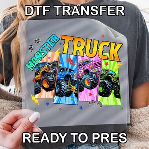 May include: A gray t-shirt with a colorful graphic of four monster trucks. The words "MONSTER TRUCK" are in bold, outlined letters. The text "DTF TRANSFER" and "READY TO PRES" are also visible.
