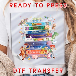 May include: DTF transfer featuring a stack of colorful VHS tapes with Disney movie titles and characters. The words "READY TO PRESS" and "DTF TRANSFER" are printed in red. The design includes characters from Toy Story, The Lion King, and more.