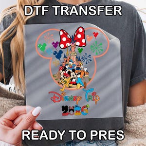 May include: A DTF transfer featuring a Disney Trip 2026 design. The design includes Mickey Mouse ears, a castle, and various Disney characters. The words "DTF TRANSFER" and "READY TO PRESS" are printed at the top and bottom.