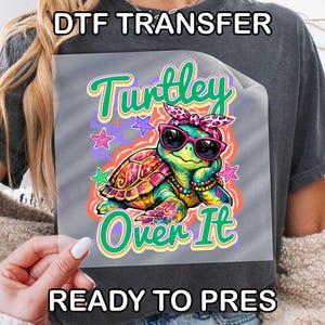 May include: A DTF transfer sheet featuring a colorful cartoon turtle wearing pink sunglasses and a leopard print headband. The turtle is surrounded by small stars and the text "Turtley Over It" in green and yellow letters. The design is on a clear film.