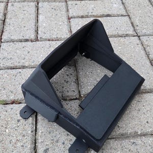 Può includere: Un paraluce per monitor in metallo nero opaco con cornice rettangolare e visiera superiore inclinata. La struttura presenta pannelli laterali, un'apertura centrale e quattro piccole alette di montaggio con fori circolari alla base.