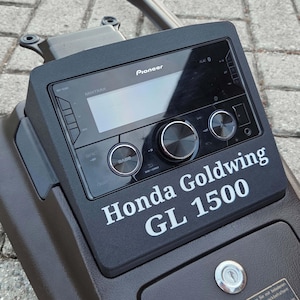 以下が含まれることがあります： ホンダ ゴールドウイング GL 1500用のダークブラウンのコンソールに設置された黒いパイオニア製カーオーディオヘッドユニット。デジタルディスプレイ、2つの円形コントロールノブ、コンソールに白でプリントされた「Honda Goldwing GL 1500」の文字が特徴です。