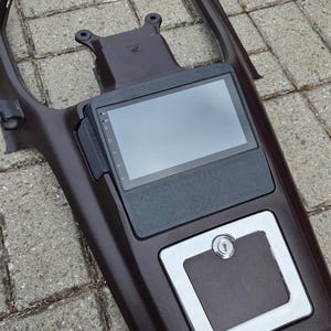 May include: A dark brown car console featuring an integrated touchscreen display, a small storage area, and a chrome-edged panel with a keyhole. The console is constructed from a dark brown material with a smooth surface.