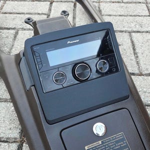 Peut inclure: Une console centrale automobile marron foncé équipée d'un autoradio Pioneer Mixtrax noir avec écran numérique, trois boutons de commande circulaires et un compartiment de rangement rectangulaire avec serrure à clé et étiquette d'avertissement.