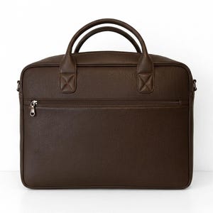 May include: A dark brown briefcase with a textured surface, featuring two rounded top handles, a horizontal zippered front pocket with a silver-toned metal pull, and side attachments for a shoulder strap, presented against a plain white background.