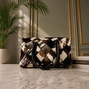 May include: A rectangular handbag featuring a patchwork design of cowhide squares in shades of brown, black, and white. The bag has two black leather handles attached to the top, with a visible quilted texture across the entire exterior surface.