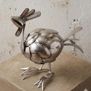May include: A handcrafted metal rooster sculpture made from repurposed silverware. The body is constructed from spoons, the head and tail from forks, and the legs from forks. The sculpture is a unique piece of art, perfect for kitchen or dining room decor.