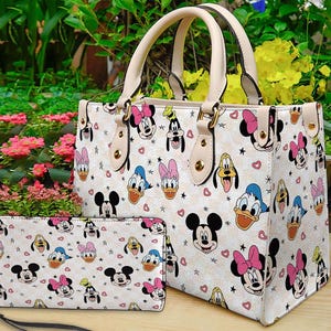 Puede incluir: Un bolso tote de color crema y una cartera a juego con un patr&oacute;n repetido de caras de personajes de dibujos animados, corazones y estrellas. El bolso tiene asas y ribetes de color crema. La cartera tiene una correa negra.
