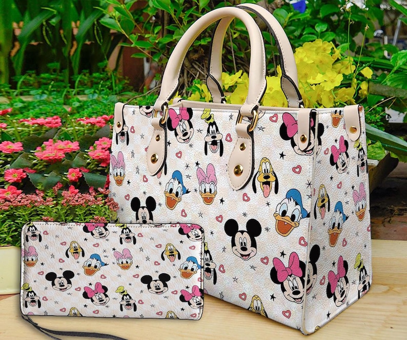 Puede incluir: Un bolso tote de color crema y una cartera a juego con un patr&oacute;n repetido de caras de personajes de dibujos animados, corazones y estrellas. El bolso tiene asas y ribetes de color crema. La cartera tiene una correa negra.