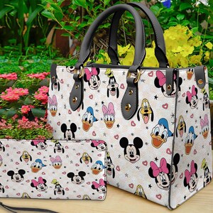 Puede incluir: Un bolso de mano blanco y una cartera a juego con un patr&oacute;n repetido de personajes de dibujos animados, incluyendo Mickey Mouse, Minnie Mouse, Donald Duck y Pluto. El bolso tiene asas negras y herrajes dorados. La cartera tiene una correa de mu&ntilde;eca.