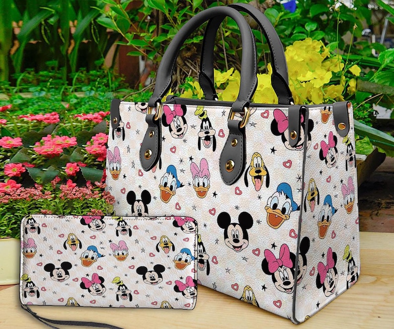 Puede incluir: Un bolso de mano blanco y una cartera a juego con un patr&oacute;n repetido de personajes de dibujos animados, incluyendo Mickey Mouse, Minnie Mouse, Donald Duck y Pluto. El bolso tiene asas negras y herrajes dorados. La cartera tiene una correa de mu&ntilde;eca.