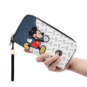 May include: A zip-around wallet featuring a denim-like blue panel with a large Mickey Mouse graphic. The white panel has a repeating Mickey Mouse pattern. The wallet has a black wrist strap and a gold-colored zipper pull.