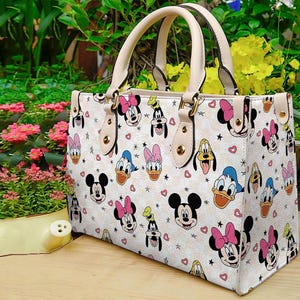 Puede incluir: Bolso tote color crema con un estampado repetido de Mickey Mouse, Minnie Mouse, Donald Duck, Daisy Duck y Pluto. El bolso tiene asas beige y herrajes dorados. Corazones y estrellas est&aacute;n dispersos por el dise&ntilde;o.