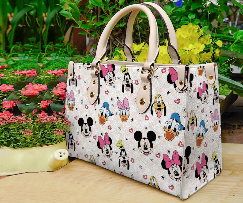 Puede incluir: Bolso tote color crema con un estampado repetido de Mickey Mouse, Minnie Mouse, Donald Duck, Daisy Duck y Pluto. El bolso tiene asas beige y herrajes dorados. Corazones y estrellas est&aacute;n dispersos por el dise&ntilde;o.
