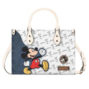 May include: White handbag featuring Mickey Mouse. The bag has a denim-like panel with Mickey Mouse, a repeating Mickey Mouse pattern, and a brown patch with the word "NAME". Includes a cream-colored handle and shoulder strap.