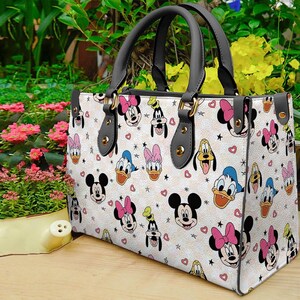 Puede incluir: Un bolso tote blanco con un patr&oacute;n repetido de caras de Mickey Mouse, Minnie Mouse, Pato Donald y Goofy. El bolso tiene asas negras y herrajes dorados. Corazones y estrellas rosas est&aacute;n dispersos por el dise&ntilde;o.