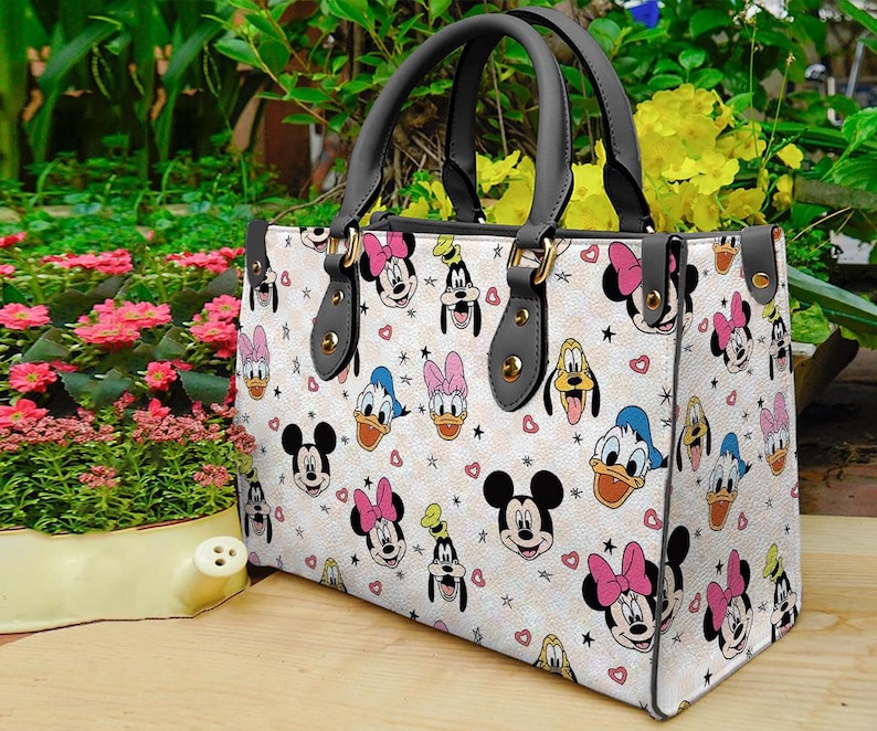 Puede incluir: Un bolso tote blanco con un patr&oacute;n repetido de caras de Mickey Mouse, Minnie Mouse, Pato Donald y Goofy. El bolso tiene asas negras y herrajes dorados. Corazones y estrellas rosas est&aacute;n dispersos por el dise&ntilde;o.
