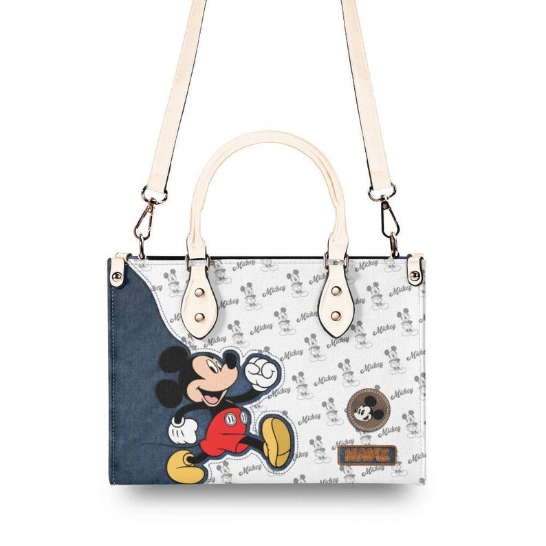 May include: White and blue handbag featuring Mickey Mouse. The bag has a white handle and a detachable shoulder strap. The design includes a large Mickey Mouse graphic and a pattern of Mickey Mouse silhouettes.