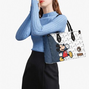 May include: White tote bag featuring a Mickey Mouse graphic and a repeating Mickey Mouse pattern. The bag has black handles and accents, with a denim-like side panel. The bag is carried by a person wearing a blue sweater and black skirt.