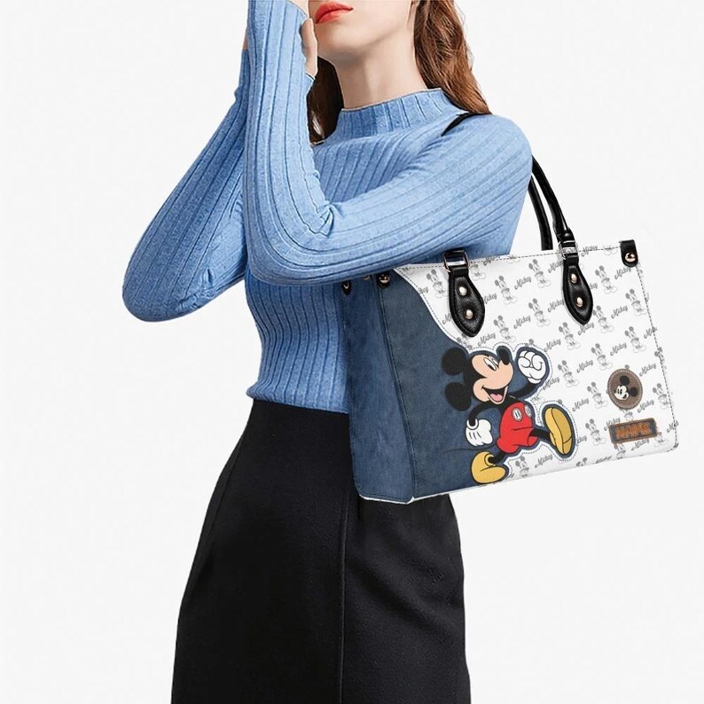 May include: White tote bag featuring a Mickey Mouse graphic and a repeating Mickey Mouse pattern. The bag has black handles and accents, with a denim-like side panel. The bag is carried by a person wearing a blue sweater and black skirt.