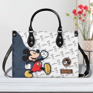 May include: White handbag featuring Mickey Mouse. The bag has a black handle and strap, with a denim-like side panel. The main body is patterned with Mickey Mouse silhouettes and the word "Mickey". A brown circular patch with Mickey's face is also visible.