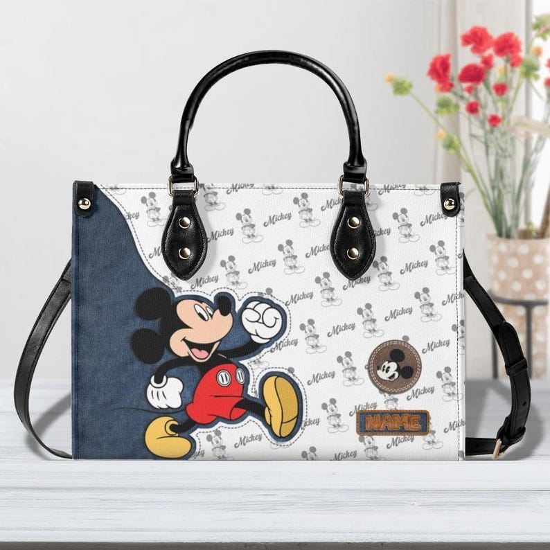 May include: White handbag featuring Mickey Mouse. The bag has a black handle and strap, with a denim-like side panel. The main body is patterned with Mickey Mouse silhouettes and the word "Mickey". A brown circular patch with Mickey's face is also visible.