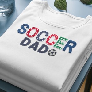 Könnte beinhalten: Ein weißes T-Shirt mit dem Text SOCCER DAD in einer mehrfarbigen Schriftart. Die Buchstaben S, O, C, C, E, R, D, A, D sind mit verschiedenen Mustern gefüllt, darunter Sterne, Punkte und Karos. Ein kleines schwarz-weißes Fußball-Symbol ist enthalten.