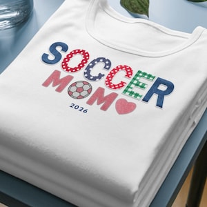 Puede incluir: Una camiseta blanca con el texto SOCCER MOM en una fuente con varios patrones de estrellas rojas, azul y cuadros verdes. La letra O de soccer es un gráfico de balón de fútbol y un corazón rosa se sitúa debajo de la palabra MOM con el año 2026.