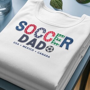 Könnte beinhalten: Ein weißes T-Shirt mit dem Schriftzug SOCCER DAD in Blau, Rot mit weißen Sternen und grünem Karomuster. Unter dem Text sind die Wörter USA, MEXICO, CANADA in Blau sowie eine kleine Grafik eines schwarz-weißen Fußballs abgebildet.