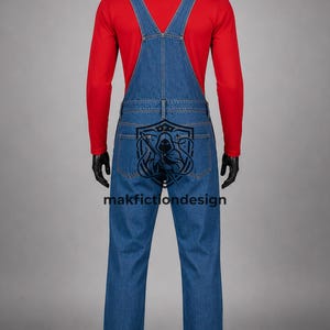 Puede incluir: Una camiseta roja de manga larga debajo de un peto vaquero azul. La parte trasera del peto presenta un estampado gr&aacute;fico negro de una figura con capucha dentro de un emblema de escudo, con el texto makfictiondesign impreso en negro.