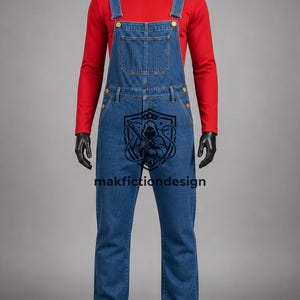 Puede incluir: Una camiseta roja de manga larga bajo un peto vaquero azul con botones dorados y un bolsillo en el pecho. En la parte delantera hay un gr&aacute;fico negro de una figura encapuchada dentro de un escudo junto al texto makfictiondesign.