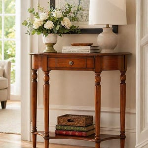 May include: A wooden console table with a semi-circular top, a single central drawer featuring a round knob, and four turned legs. The table includes a lower shelf holding a woven basket and two books, with a lamp and flowers on top.