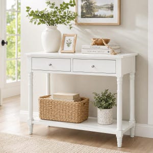 May include: A white console table with two drawers, a lower shelf, and turned legs. The table is decorated with a white vase with greenery, a framed picture, books, a woven basket, and a potted plant. The table is approximately 36 inches wide.