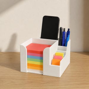 Puede incluir: Un organizador de escritorio de plástico blanco con un compartimento cuadrado grande lleno de una pila de notas adhesivas multicolores, una ranura trasera alta con un teléfono inteligente negro y tres compartimentos laterales con bolígrafos.