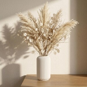 Peut inclure: Un vase cylindrique blanc à texture nervurée verticale contenant un arrangement floral séché. Le bouquet se compose de panaches d'herbe de la pampa beige duveteux et de gousses de graines de lunaire circulaires, translucides et délicates.