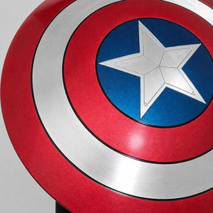 May include: A circular shield featuring a metallic red outer ring, a silver middle ring, and a central blue circle containing a large, five-pointed silver star. The design is symmetrical with distinct color bands and a smooth, polished finish.