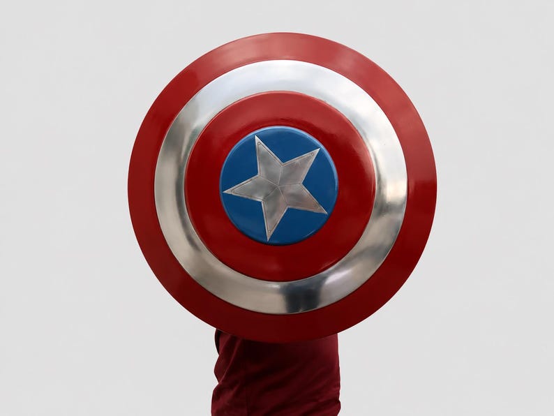 May include: A circular shield featuring a concentric design with a bright red outer ring, a metallic silver middle ring, and a blue center circle containing a large, five-pointed silver star. The shield has a smooth, reflective finish.