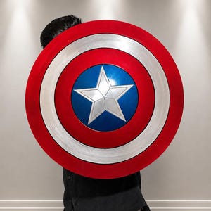 May include: A circular shield featuring a central silver star on a blue background, surrounded by a white ring and an outer red ring. The design includes concentric bands of metallic silver, red, and blue, creating a classic patriotic appearance.