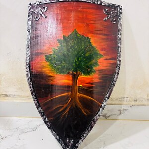 May include: A heater-style shield featuring a hand-painted scene of a green tree with visible roots set against a vibrant orange and red sunset sky. The shield has a metallic silver-coloured border with a distressed finish and decorative accents.