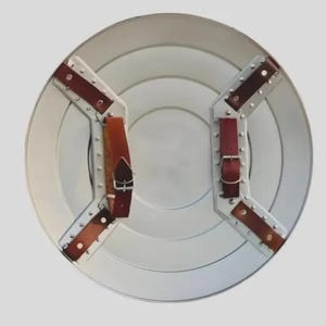 May include: A circular silver-toned metal shield viewed from the back. It features two white metal mounting brackets secured with rivets, each holding brown leather straps with silver-colored metal buckles for adjustable attachment.