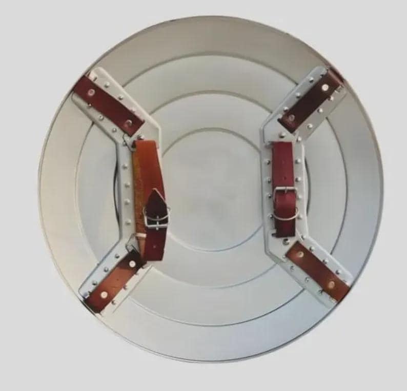 May include: A circular silver-toned metal shield viewed from the back. It features two white metal mounting brackets secured with rivets, each holding brown leather straps with silver-colored metal buckles for adjustable attachment.