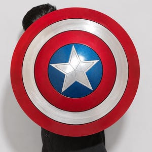 May include: A circular shield featuring a metallic silver star centered on a blue circular background. The design includes concentric rings of silver and red, with a brushed metal texture on the silver sections and a smooth finish on the red.