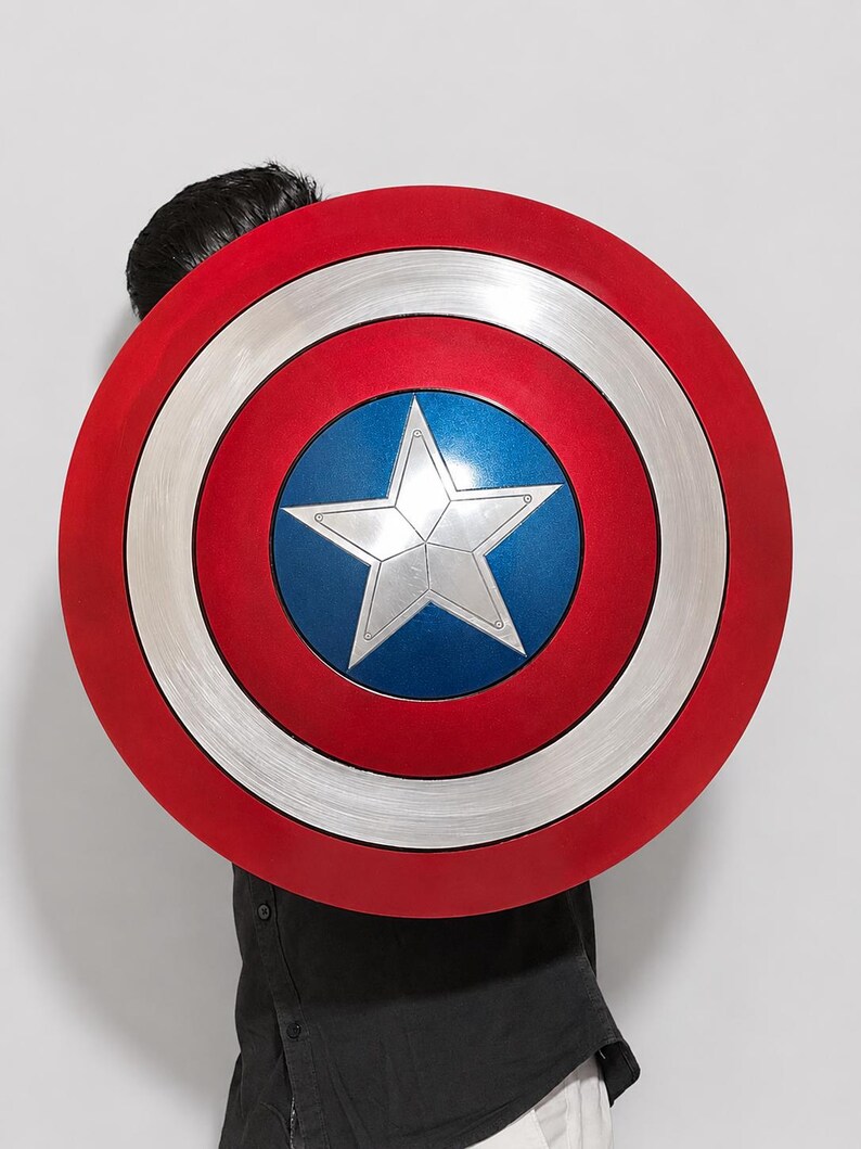 May include: A circular shield featuring a metallic silver star centered on a blue circular background. The design includes concentric rings of silver and red, with a brushed metal texture on the silver sections and a smooth finish on the red.